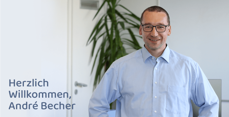 Offizielles Begrüßungsbild für André Becher in einem Büroumfeld. Es zeigt den Mitarbeiter lächelnd mit Brille und blauem Hemd vor einer weißen Wand mit einer Pflanze. Der Schriftzug „Herzlich Willkommen, André Becher“ ist in der unteren linken Ecke platziert.