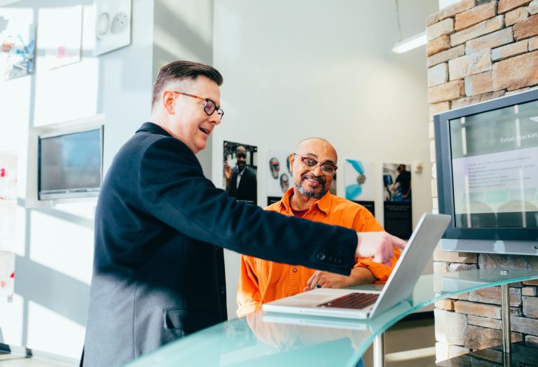 Zwei Geschäftsleute stehen in einem modernen Büro an einem Glastresen und betrachten gemeinsam einen Laptop. Ein Mann im dunklen Anzug und mit Brille zeigt mit dem Finger auf den Bildschirm, während sein Kollege im orangefarbenen Hemd lächelnd danebensteht. Im Hintergrund sind ein heller Raum mit einer Steinwand und Monitore zu sehen.