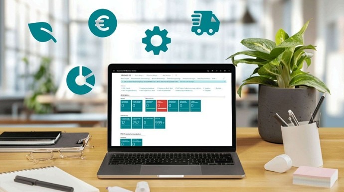 Ein Laptop auf einem Schreibtisch in einem hellen Büro, auf dessen Bildschirm eine ERP-Software (Microsoft Dynamics 365 Business Central) mit Dashboard-Kacheln und Menüs geöffnet ist. Über dem Laptop schweben verschiedene türkisfarbene Symbole wie ein Blatt, ein Euro-Zeichen, ein Zahnrad, ein Lkw und ein Diagramm. Rechts daneben stehen eine Topfpflanze und ein Stiftehalter.