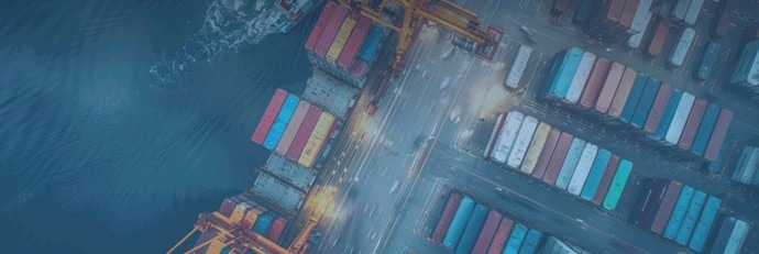 Eine in Blautöne getauchte Luftaufnahme eines geschäftigen Containerhafens bei Nacht oder Dämmerung. Zu sehen sind ein Frachtschiff an der Kaikante, Hafenkräne sowie ordentlich aufgereihte, bunte Frachtcontainer an Land.
