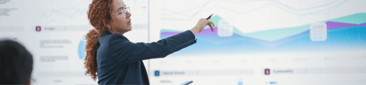 Eine Frau mit Brille und lockigen Haaren in einem dunklen Blazer hält eine Präsentation vor einer Leinwand oder einem Whiteboard, auf dem bunte Diagramme und Analysen zu sehen sind, und zeigt mit einem Stift auf eine Grafik.