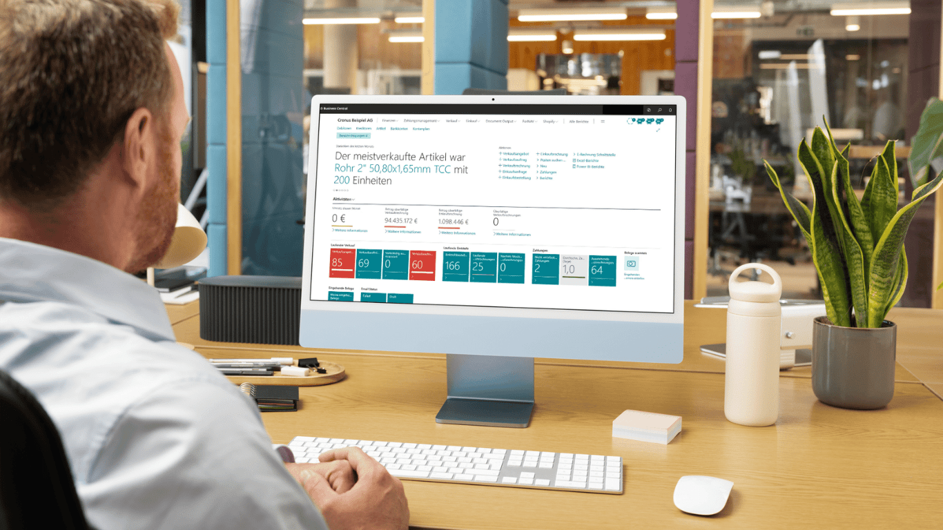 Ein Benutzer sitzt an einem Schreibtisch und betrachtet einen Monitor, auf dem das Business Central Dashboard angezeigt wird. Das Dashboard zeigt eine Projektübersicht mit einem verkauften Artikel "Rohr 2" sowie verschiedene farbige KPI-Kacheln mit Kennzahlen wie Umsatz (94.435 172 €), Soll-Wert (1.088 446 €) und weitere Metriken zur Projektverfolgung.
